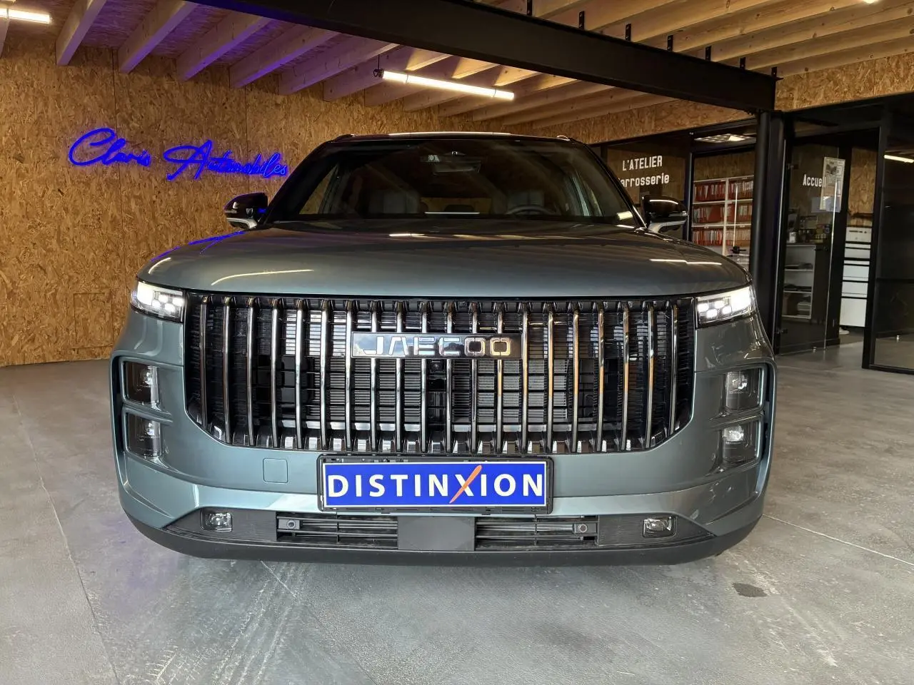 Vue frontale du SUV JAECOO J7 2026 en gris olive avec calandre verticale noire et toit noir dans un showroom.