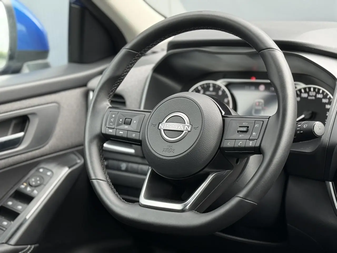 Vue intérieure centrée sur le volant noir du Nissan Qashqai 2025, avec portière côté gauche et rétroviseur bleu visible.