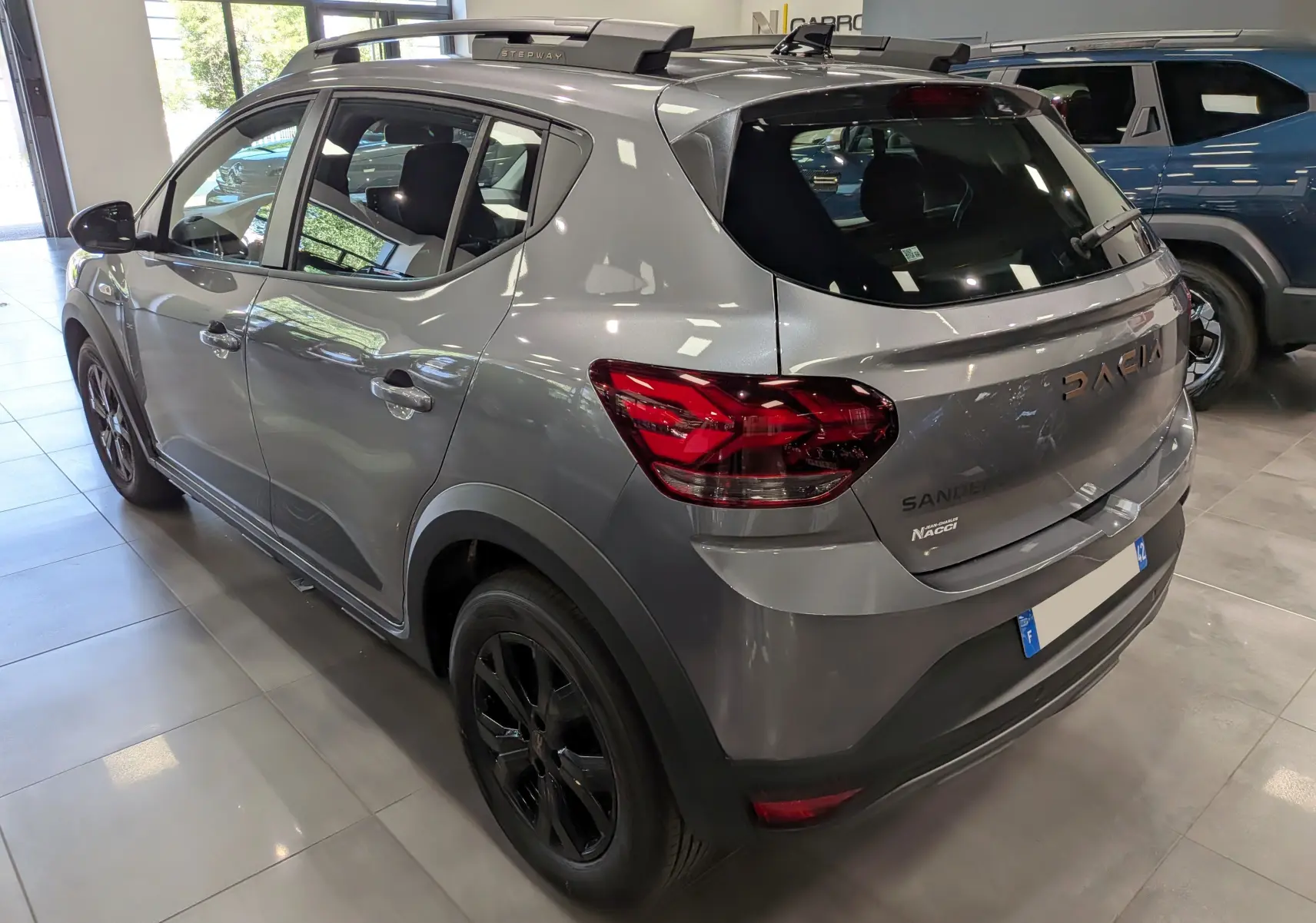 Vue 3/4 arrière droite d’une Dacia Sandero Stepway gris schiste avec jantes noires et barres de toit en concession.