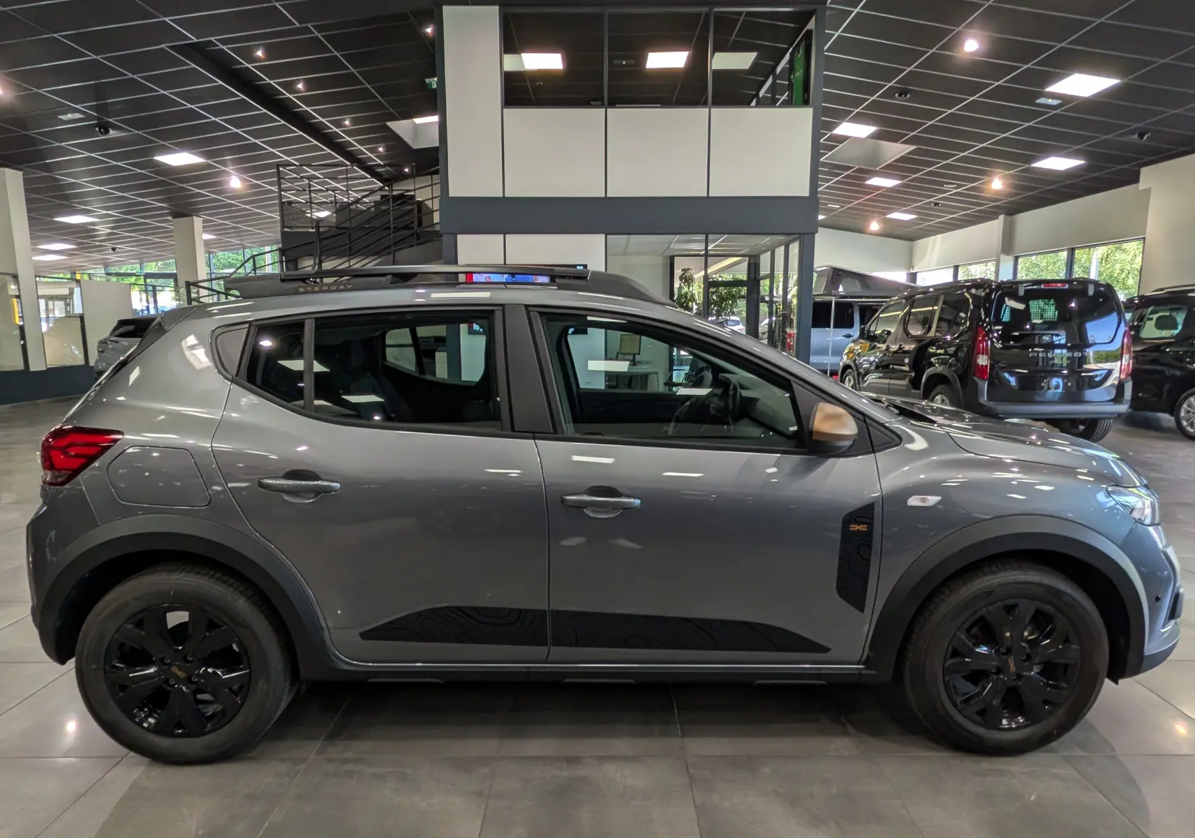 Vue de profil droit d'une Dacia Sandero Stepway TCe 110 gris schiste avec jantes noires en intérieur de showroom.