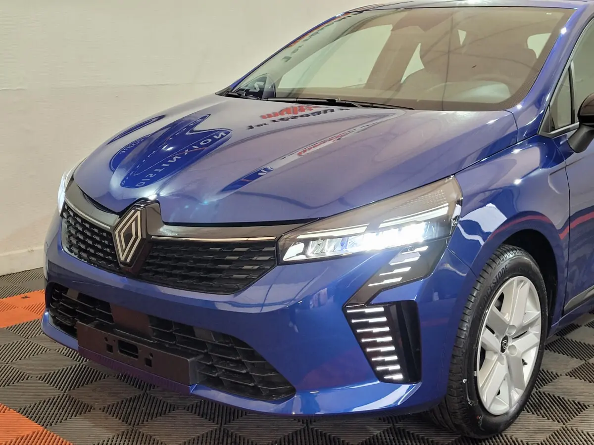 Vue 3/4 avant droite d'une Renault Clio bleu avec phares LED allumés et jantes argentées visibles.