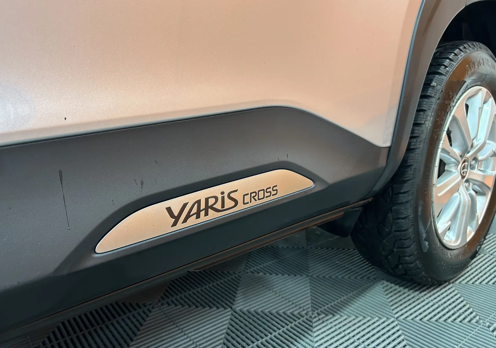 Gros plan sur la partie basse côté droit d'une Toyota Yaris Cross gris minéral avec logo Yaris Cross et roue arrière visible.