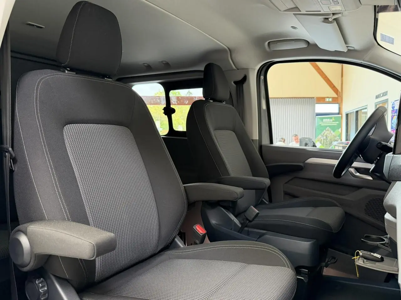 Vue intérieure côté gauche du Volkswagen Transporter T7 ProCab 2026, sièges tissu gris à rayures avec accoudoirs.