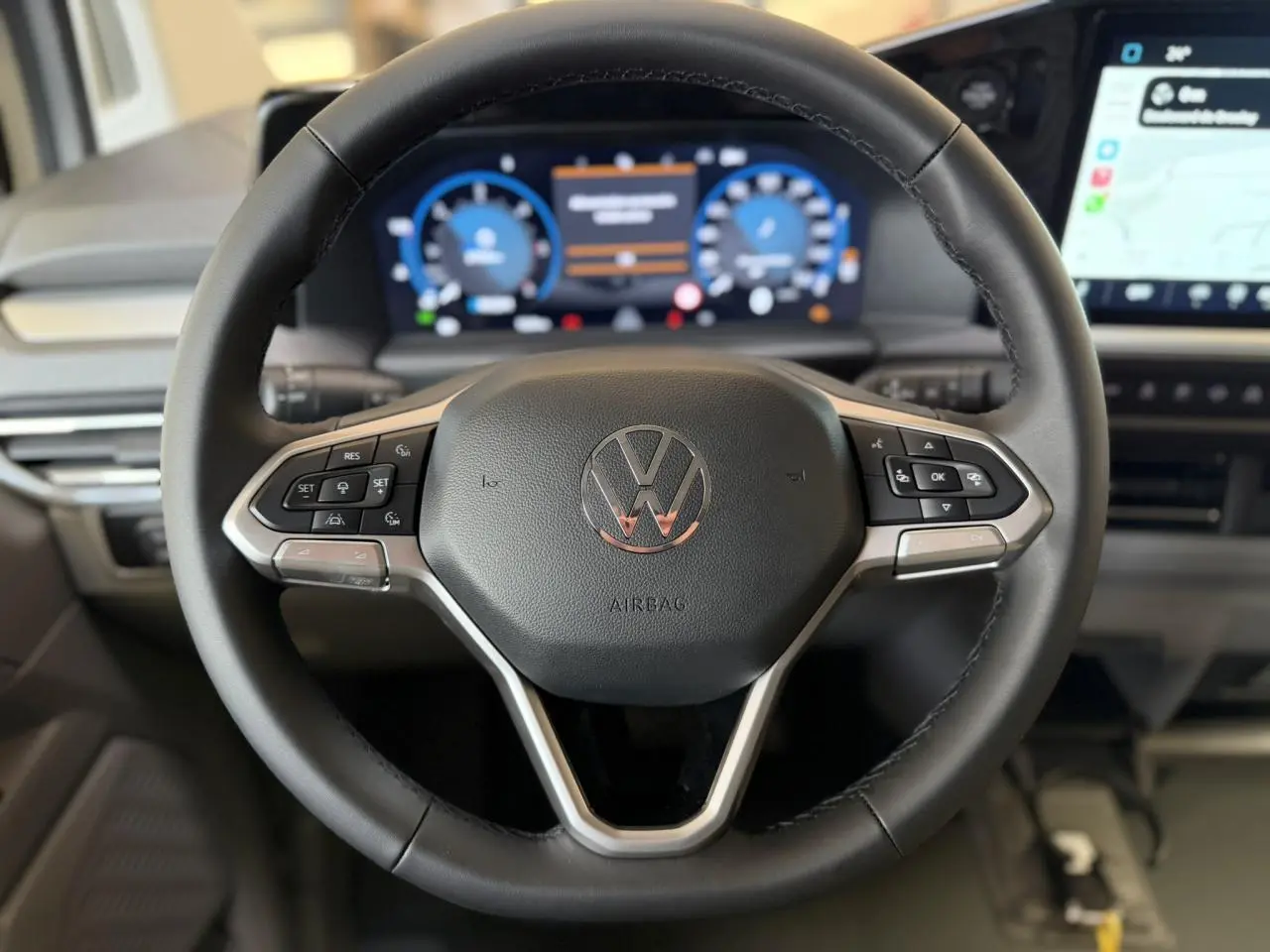 Volant multifonction noir du Volkswagen Transporter T7 avec tableau de bord numérique et écran tactile visible en arrière-plan.