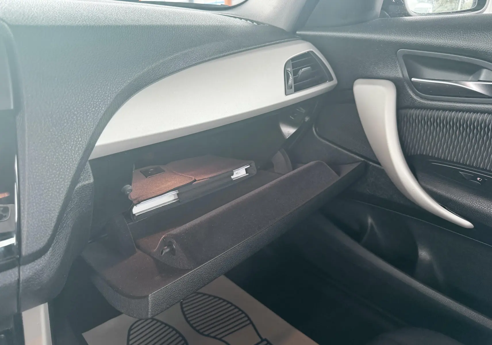 Intérieur BMW Série 1 2016, vue côté passager sur la boîte à gants ouverte avec manuel visible, garnitures noires et grises.