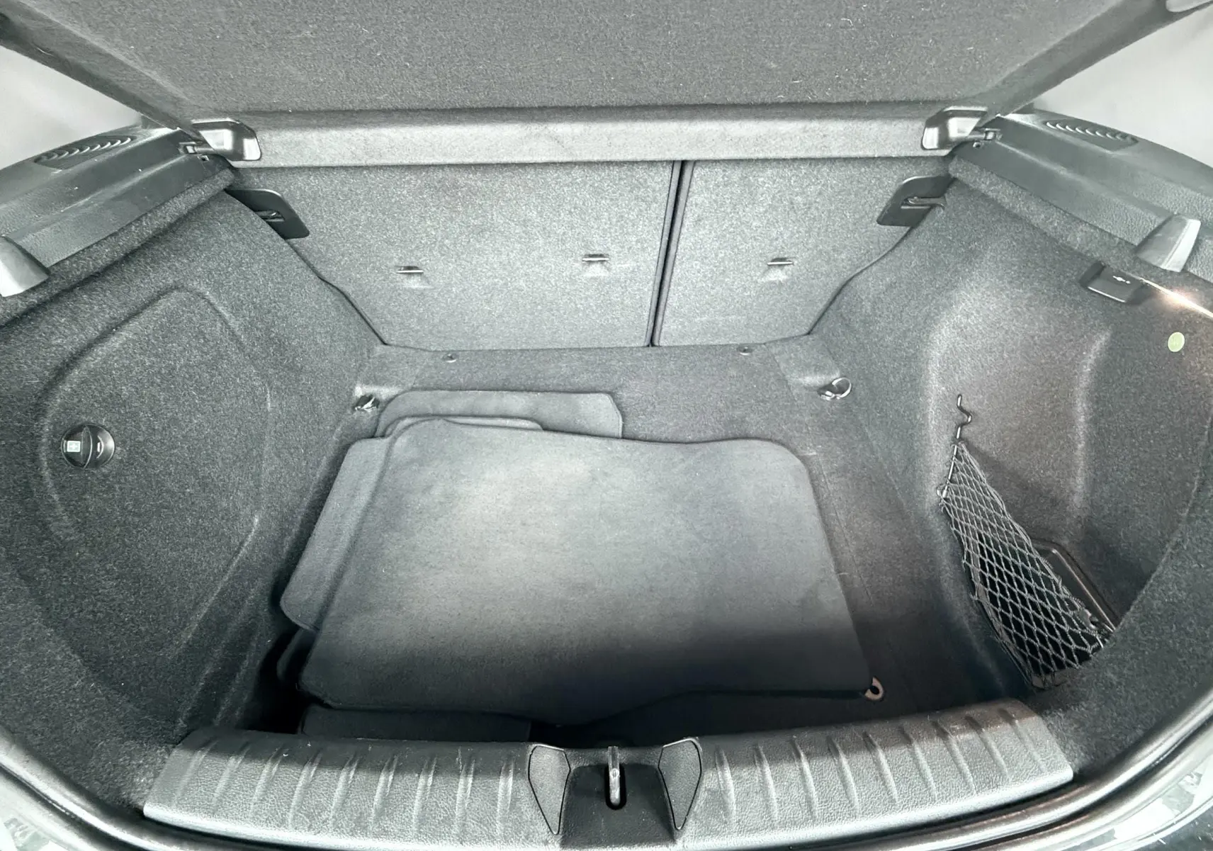 Coffre ouvert de BMW Série 1 noire, vue plongeante montrant l’espace de rangement et tapis intérieur gris.