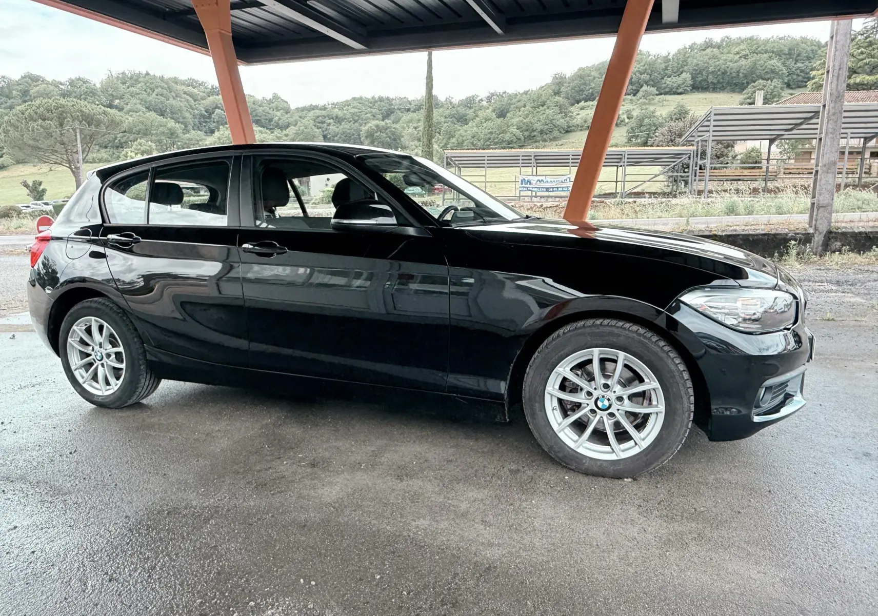 BMW Série 1 noire vue de profil côté gauche sous abri, avec jantes alliage et carrosserie brillante.