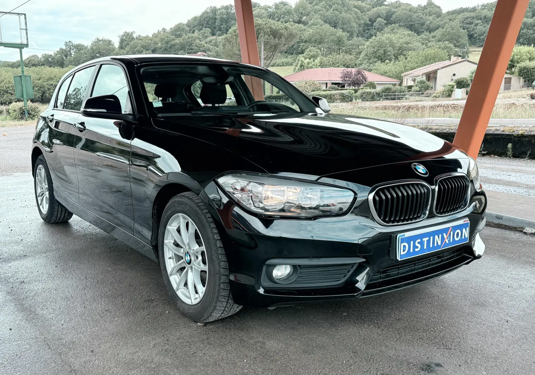 BMW Série 1 noire vue en 3/4 avant droit, sous un abri avec un paysage verdoyant en arrière-plan.