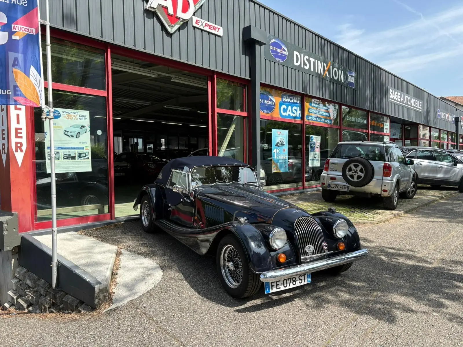 Morgan Plus 4 bleu foncé vue 3/4 avant droit, capote fermée, garée devant un garage avec façade vitrée rouge.