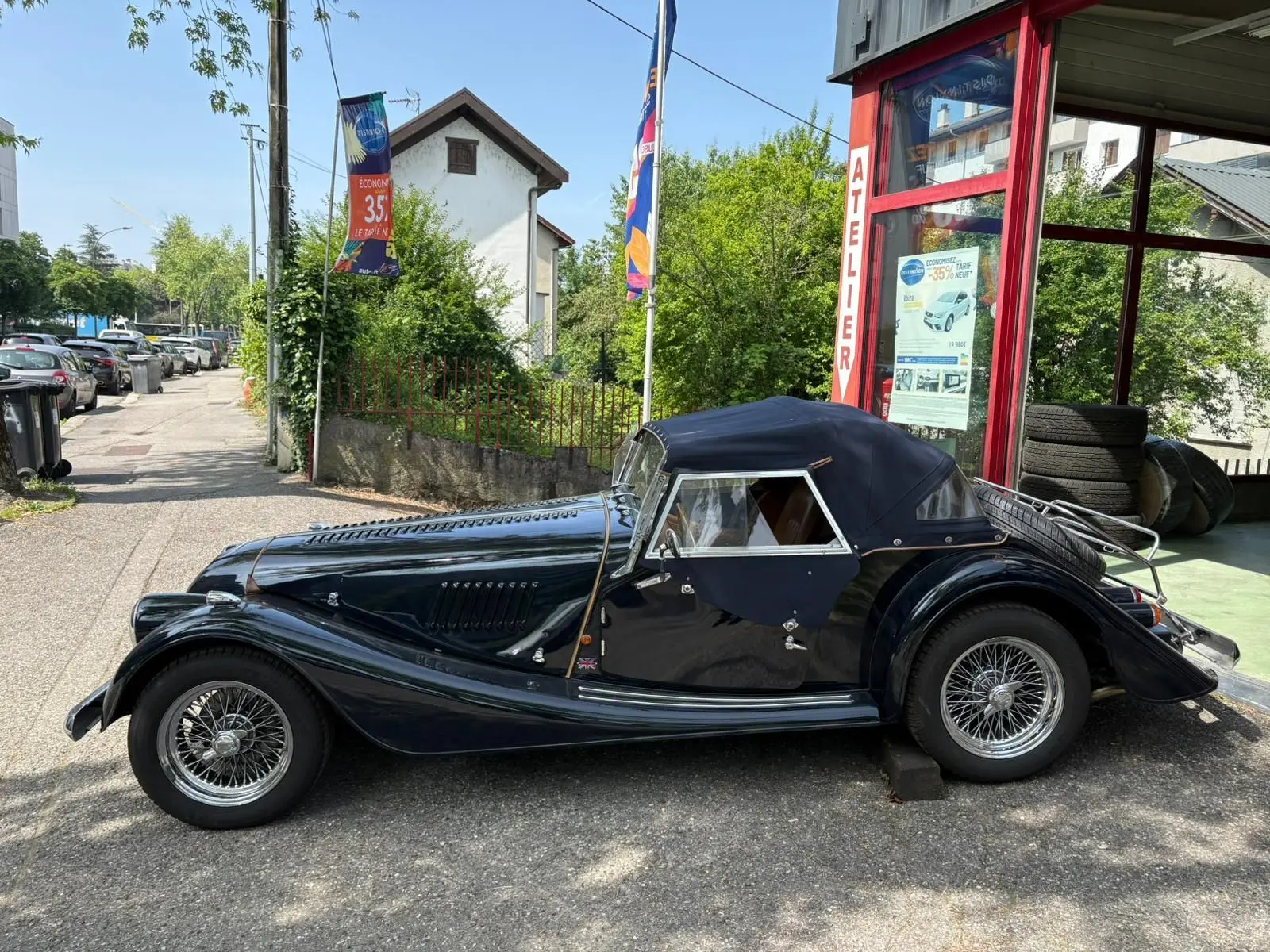Morgan Plus 4 bleu foncé vue de profil côté gauche, capote fermée et jantes à rayons chromées visibles.