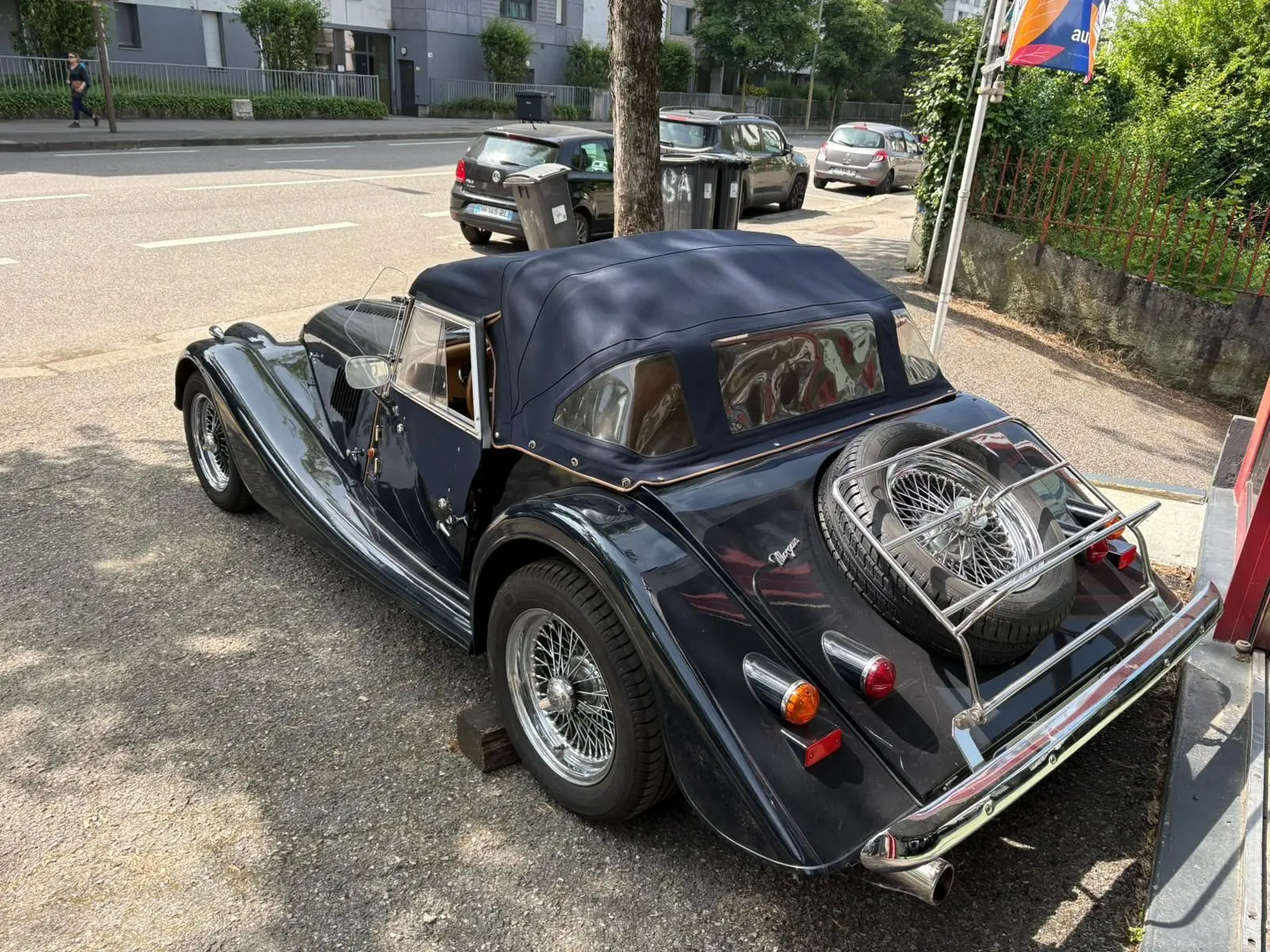 Morgan Plus 4 bleu foncé vue 3/4 arrière droit, capote fermée et roue de secours sur coffre chromé.