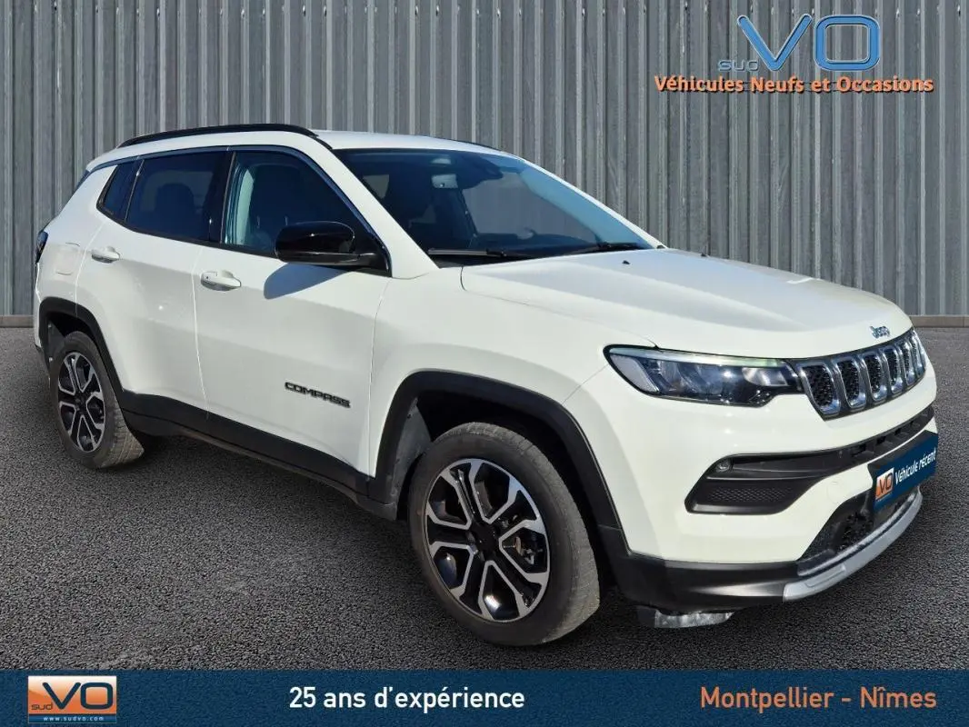 Jeep Compass blanc vue 3/4 avant droit, avec jantes 18 pouces et barres de toit noires sur fond urbain.