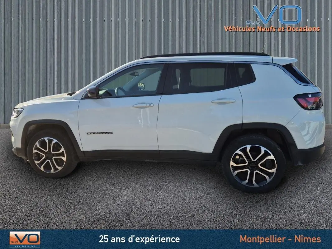 Profil latéral droit d'un Jeep Compass blanc 2023 avec jantes 18 pouces et barres de toit visibles sur fond industriel.