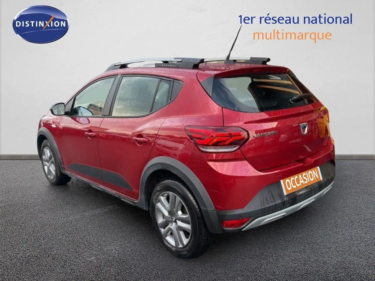 Vue 3/4 arrière droite d'une Dacia Sandero Stepway rouge fusion avec barres de toit et protections noires sur les bas de caisse.