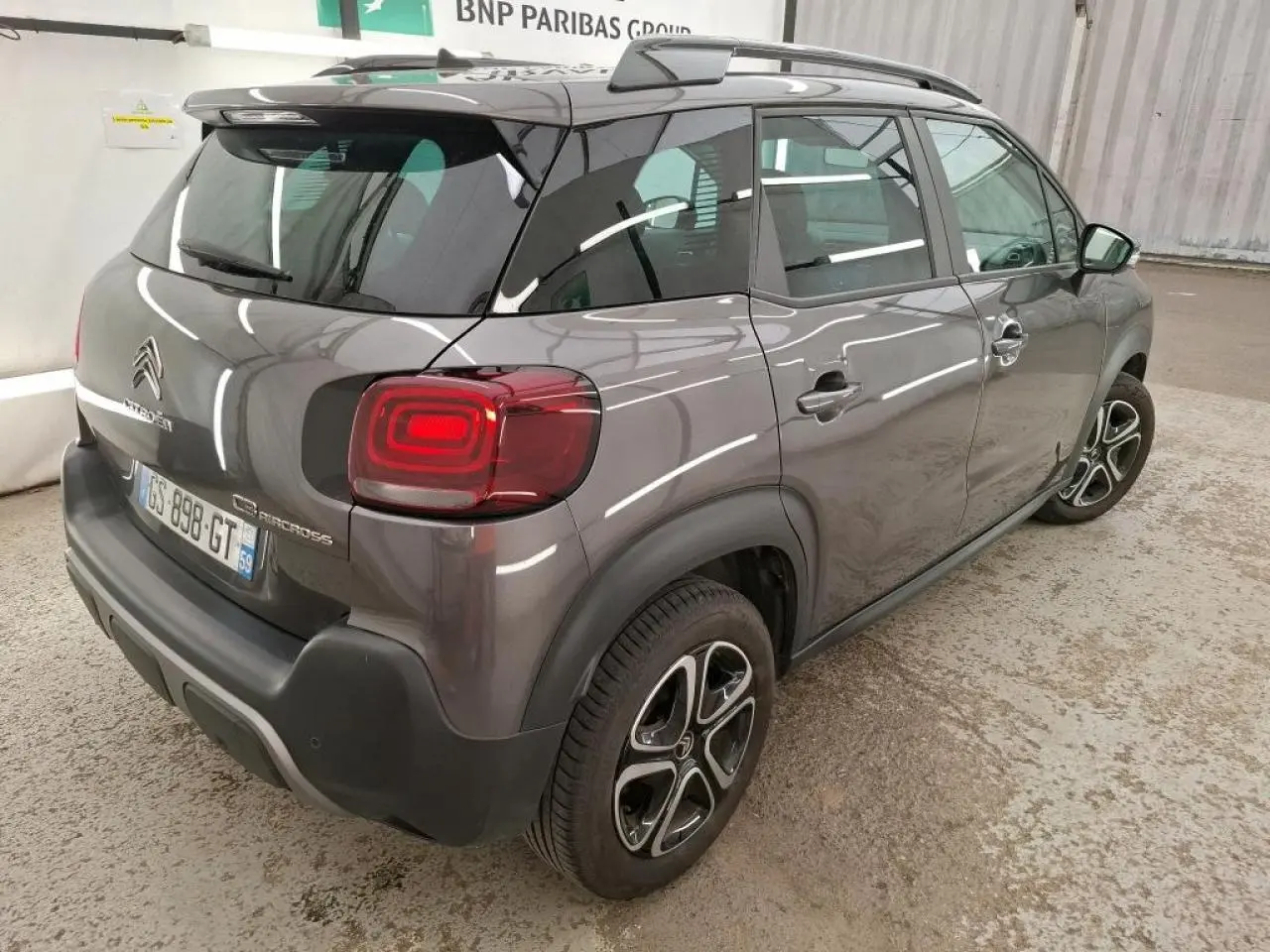 Vue 3/4 arrière droit d'un Citroën C3 Aircross gris foncé avec barres de toit noires et feux arrière LED.