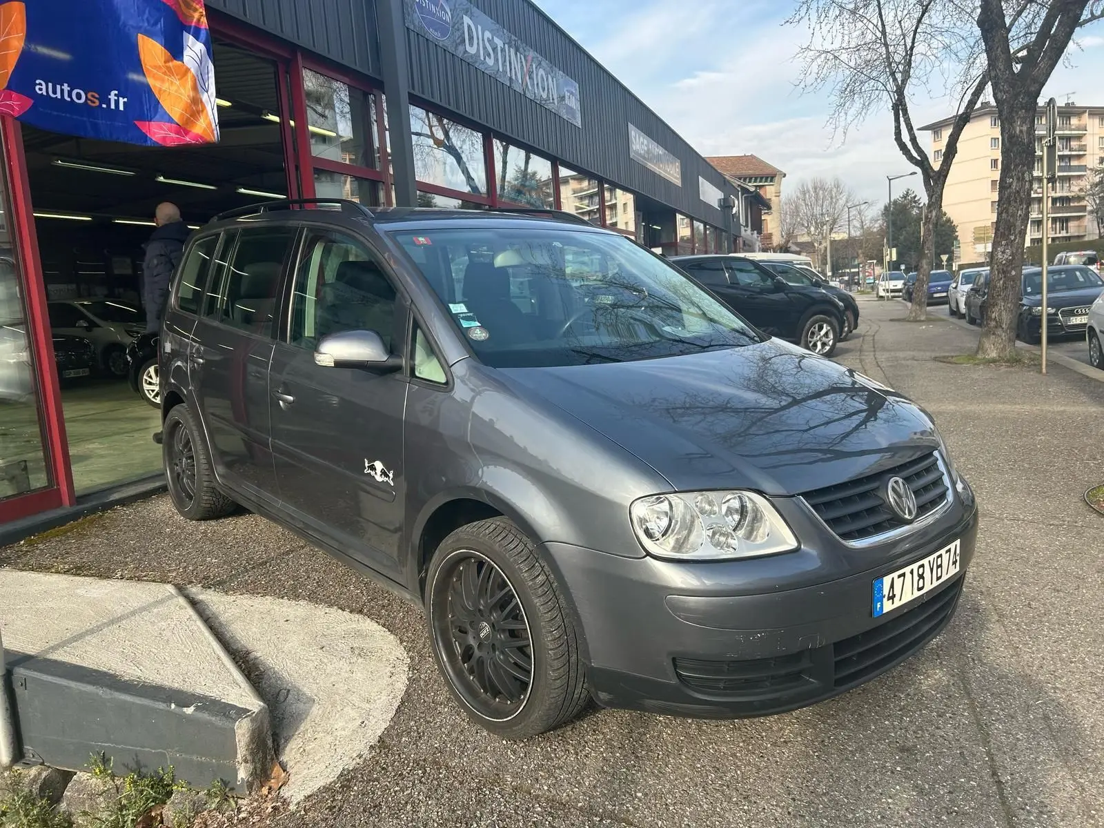 Volkswagen Touran gris vue 3/4 avant gauche avec jantes noires et sticker sur la porte côté gauche.