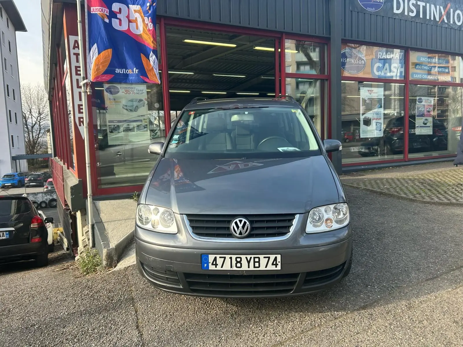Volkswagen Touran gris 2004 vu de face, stationné devant un atelier avec une bannière publicitaire visible.