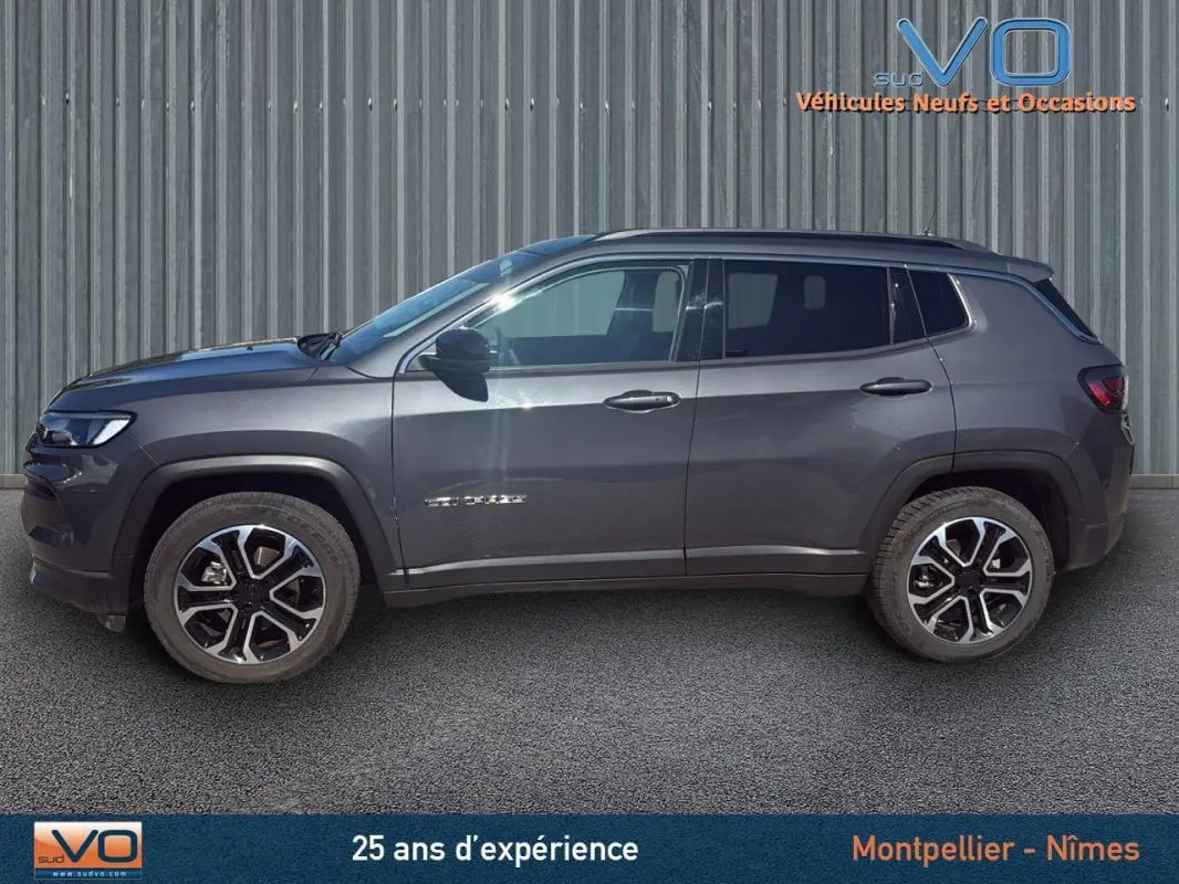 Profil côté gauche d'un Jeep Compass gris 2023 avec jantes 18 pouces et barres de toit visibles sur fond industriel.