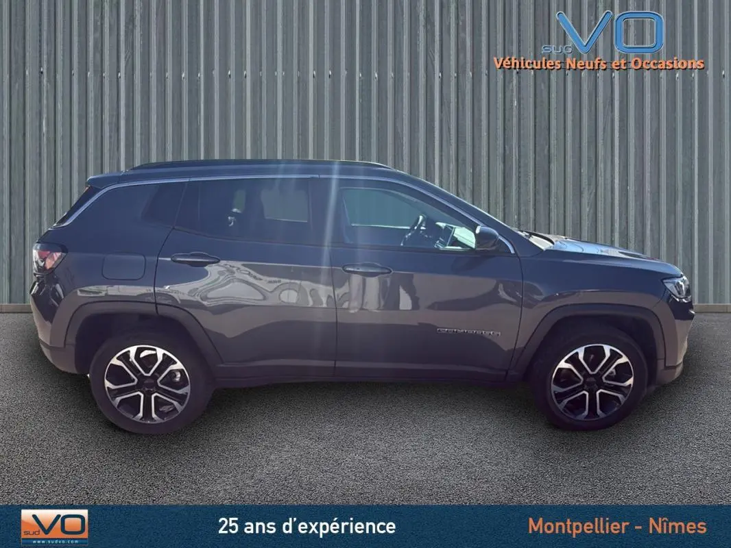 Vue latérale droite d'un Jeep Compass gris 2023 avec jantes 18 pouces et barres de toit visibles sur fond industriel.