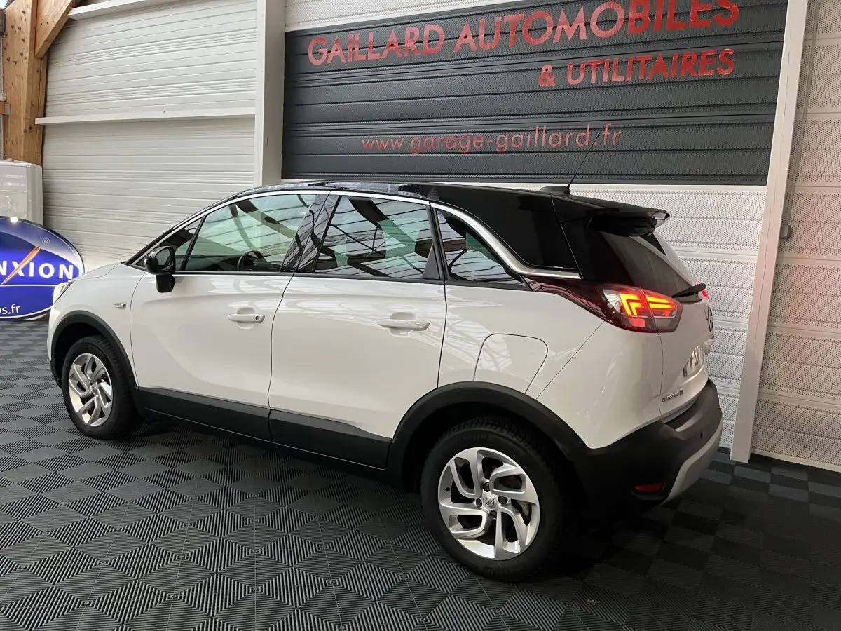 Vue 3/4 arrière droite d'un Opel Crossland X blanc avec toit noir, feux arrière allumés, stationné en intérieur.