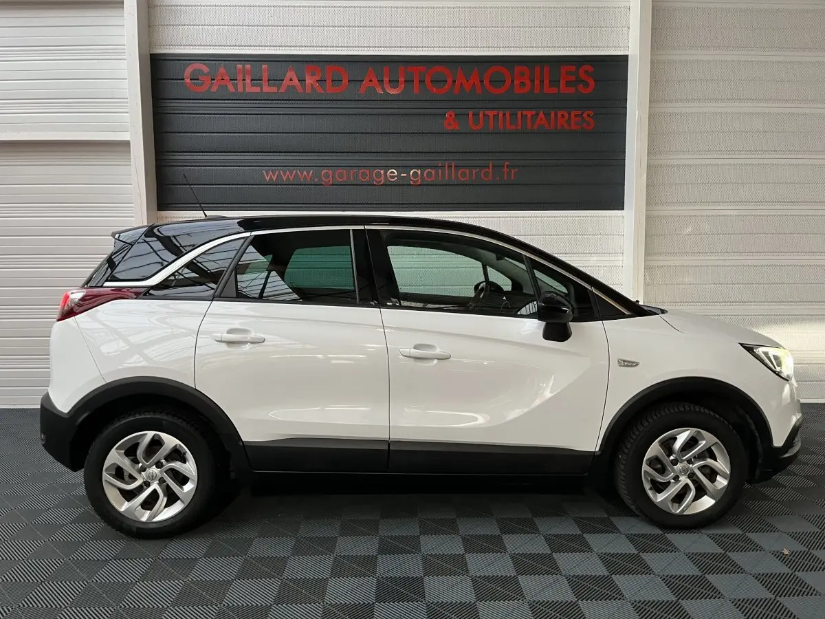 Vue de profil côté gauche d'un Opel Crossland X blanc avec toit noir, roues alliage et protections noires basses.