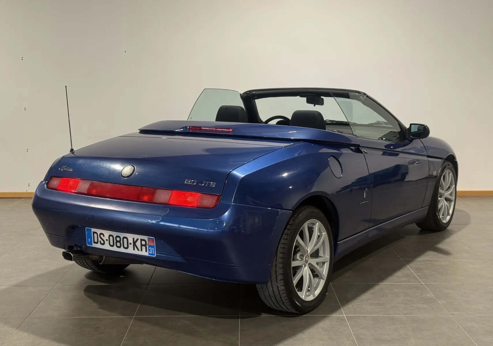 Vue 3/4 arrière droite d'un cabriolet Alfa Romeo Spider bleu 2006 avec capote ouverte et jantes argentées.