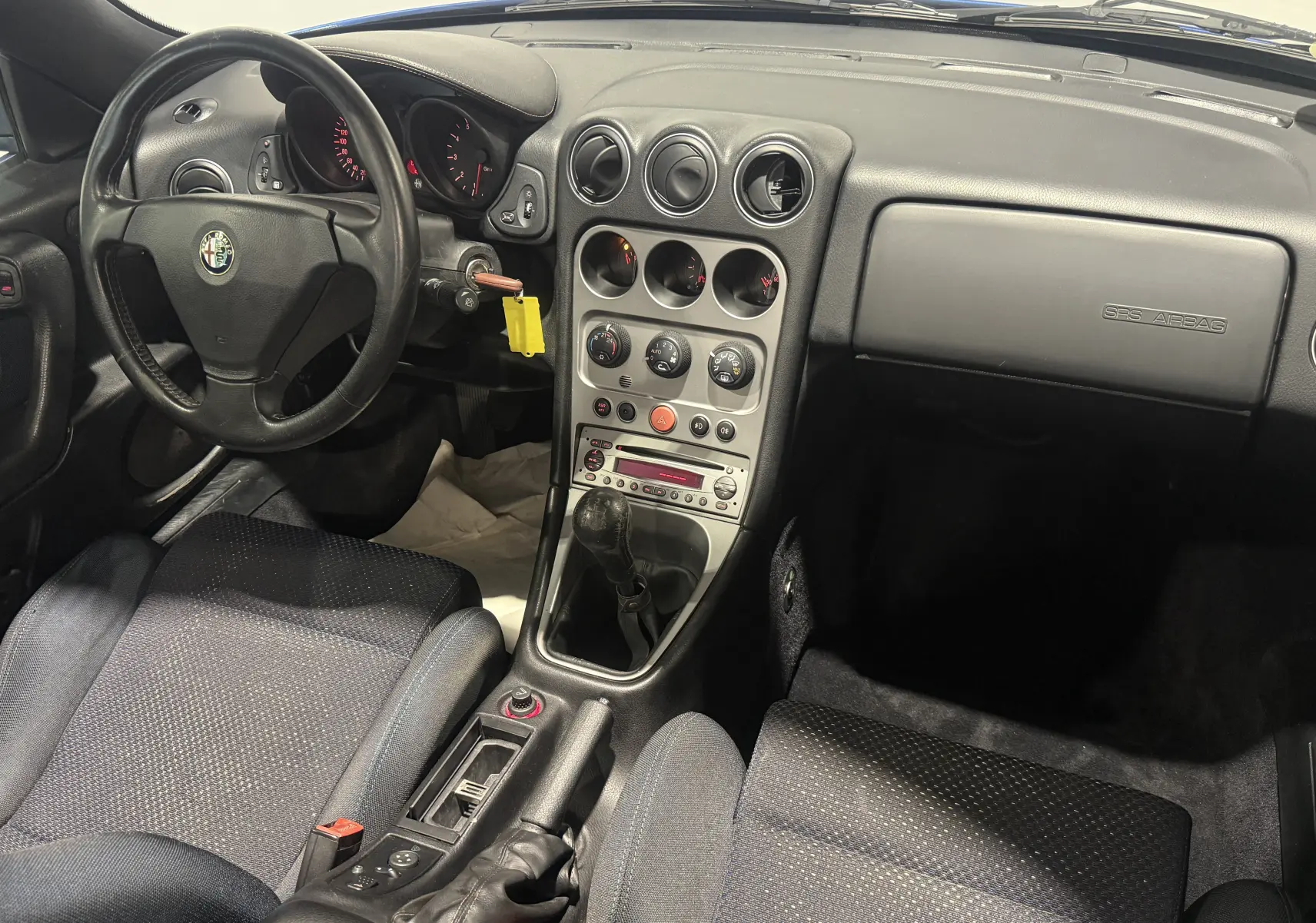 Vue intérieure côté conducteur de l'Alfa Romeo Spider bleu 2006, avec tableau de bord noir et levier de vitesses manuel.