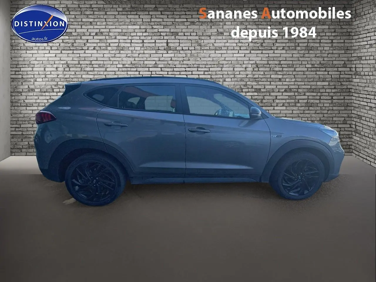 Vue latérale droite du Hyundai Tucson 1.6 Hybrid 136 N Line gris foncé avec jantes alliage noires, sur fond de mur en briques.