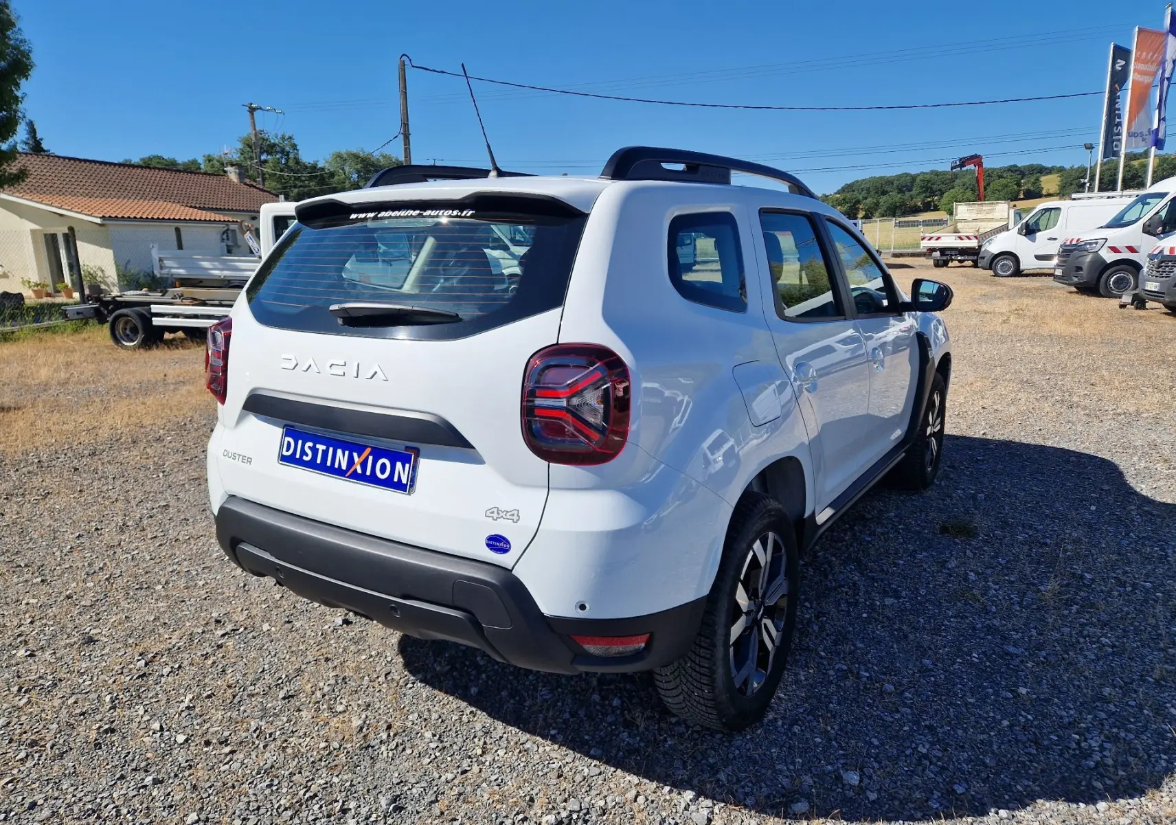 Vue 3/4 arrière droite d'un Dacia Duster blanc 4x4 2024 avec jantes alu 17 pouces sur un terrain gravillonné.
