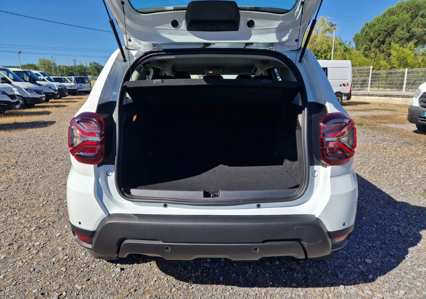 Vue arrière du coffre ouvert d'un Dacia Duster blanc 4x4 2024, avec feux arrière LED et pare-chocs noir.