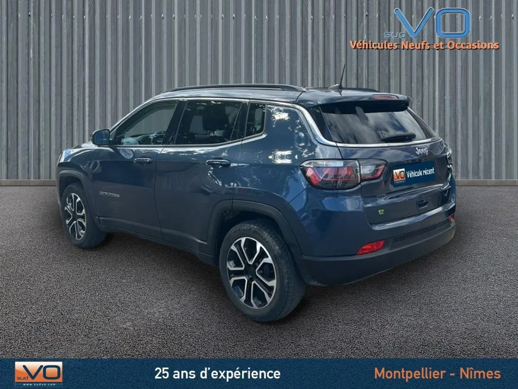 Jeep Compass bleu 3/4 arrière droit, avec barres de toit et jantes 18 pouces visibles sur fond industriel.