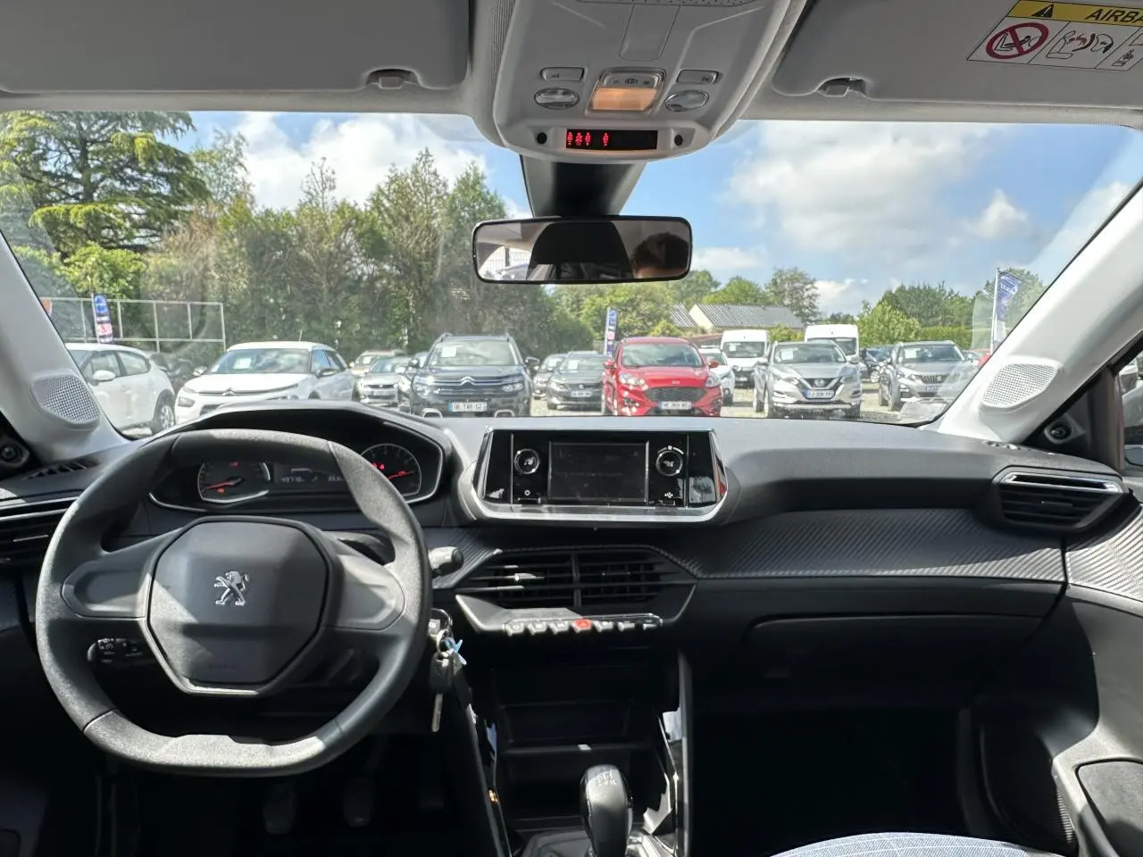 Vue intérieure avant du tableau de bord noir et volant Peugeot 208 blanc, avec écran tactile et ciel bleu visible à l'extérieur.