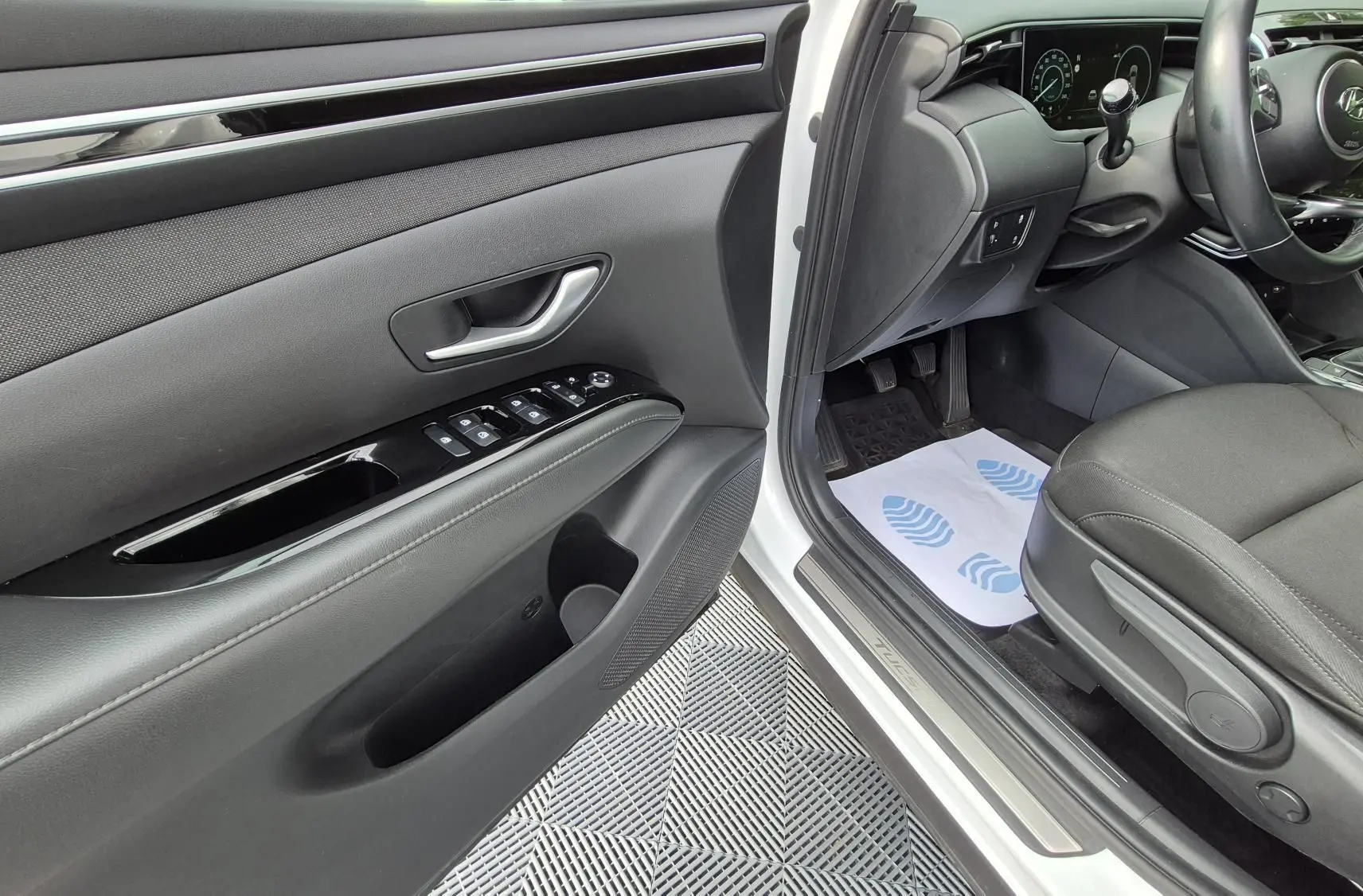 Vue intérieure côté conducteur du Hyundai Tucson blanc 2022, montrant la porte, siège tissu noir et tableau de bord.