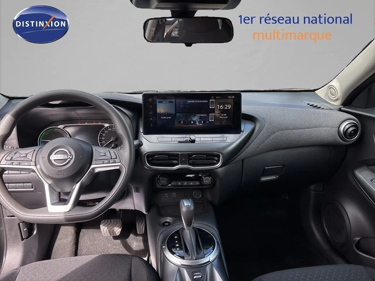 Vue intérieure frontale du tableau de bord et volant du Nissan Juke Hybrid gris, avec écran tactile central et levier de boîte automatique.