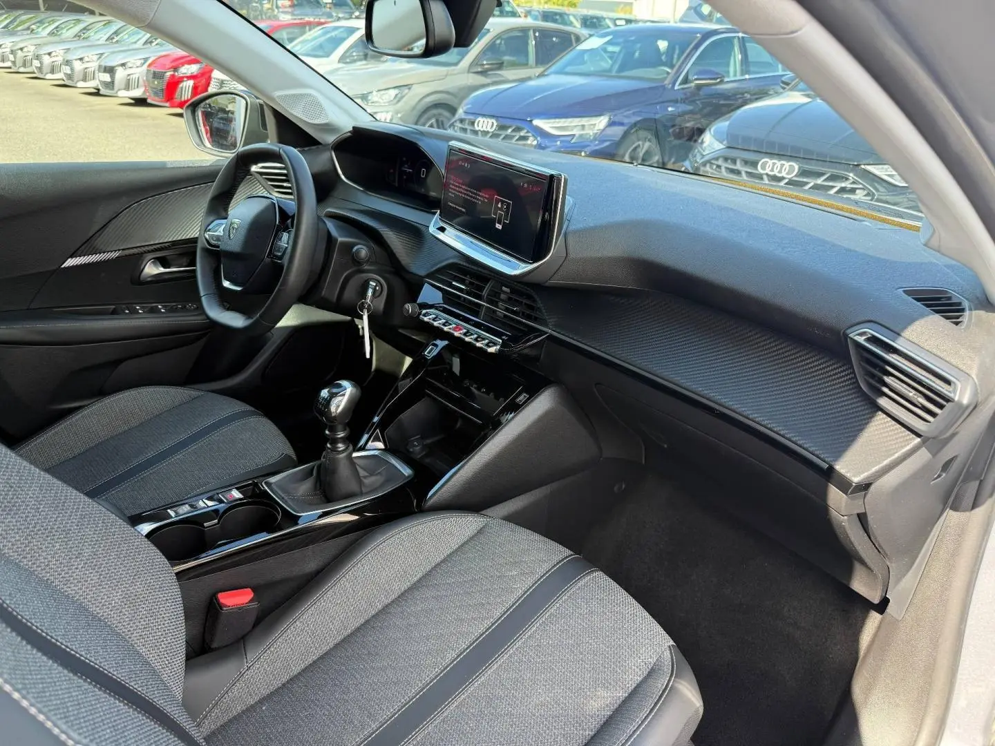 Intérieur avant droit de la Peugeot 208 1.2 100ch gris Artense, volant, écran tactile et levier de vitesse manuel visibles.