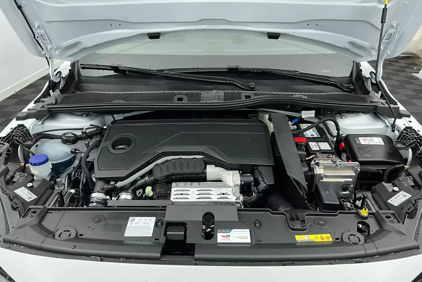 Moteur hybride Peugeot 208 1.2 HYBRID 136 GT blanc vu de face sous le capot ouvert, montrant le compartiment moteur.
