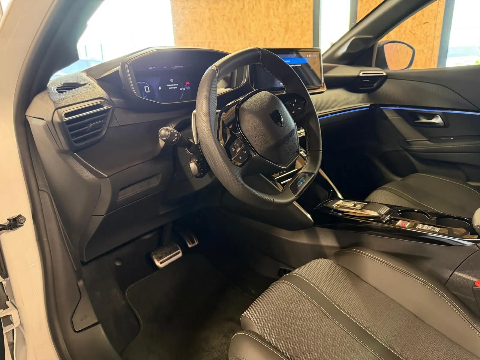 Vue intérieure côté conducteur de la Peugeot 208 1.2 Hybrid 136 GT blanche, avec volant multifonction et tableau de bord numérique.