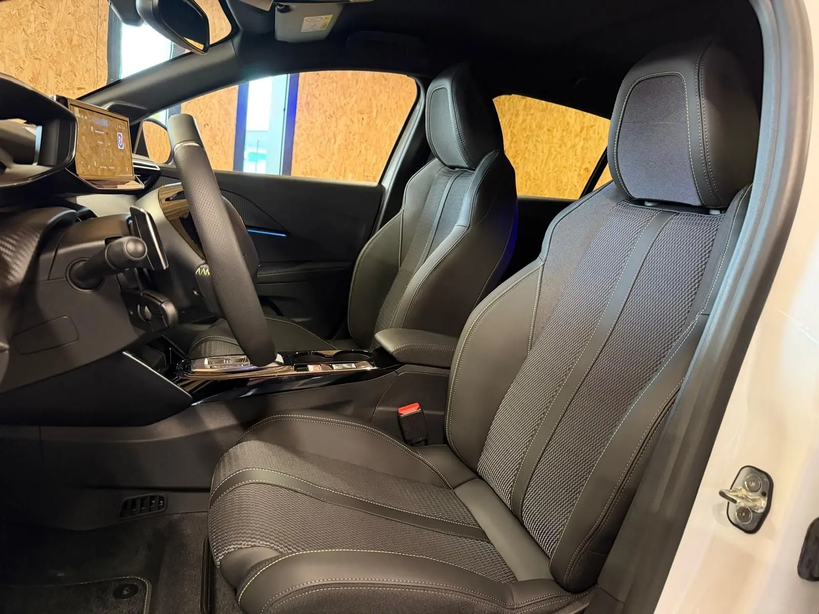 Vue intérieure côté conducteur de la Peugeot 208 blanche 2025, mettant en valeur les sièges en tissu noir et le tableau de bord moderne.