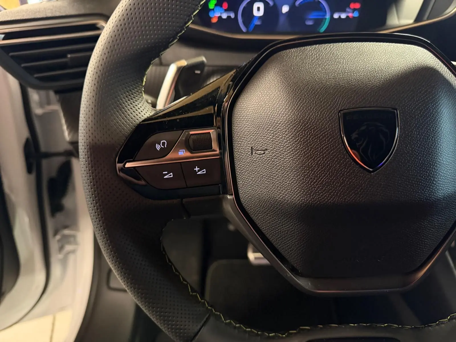 Gros plan sur le volant noir perforé du Peugeot 208 1.2 HYBRID 136 GT blanc avec commandes intégrées et tableau de bord digital.