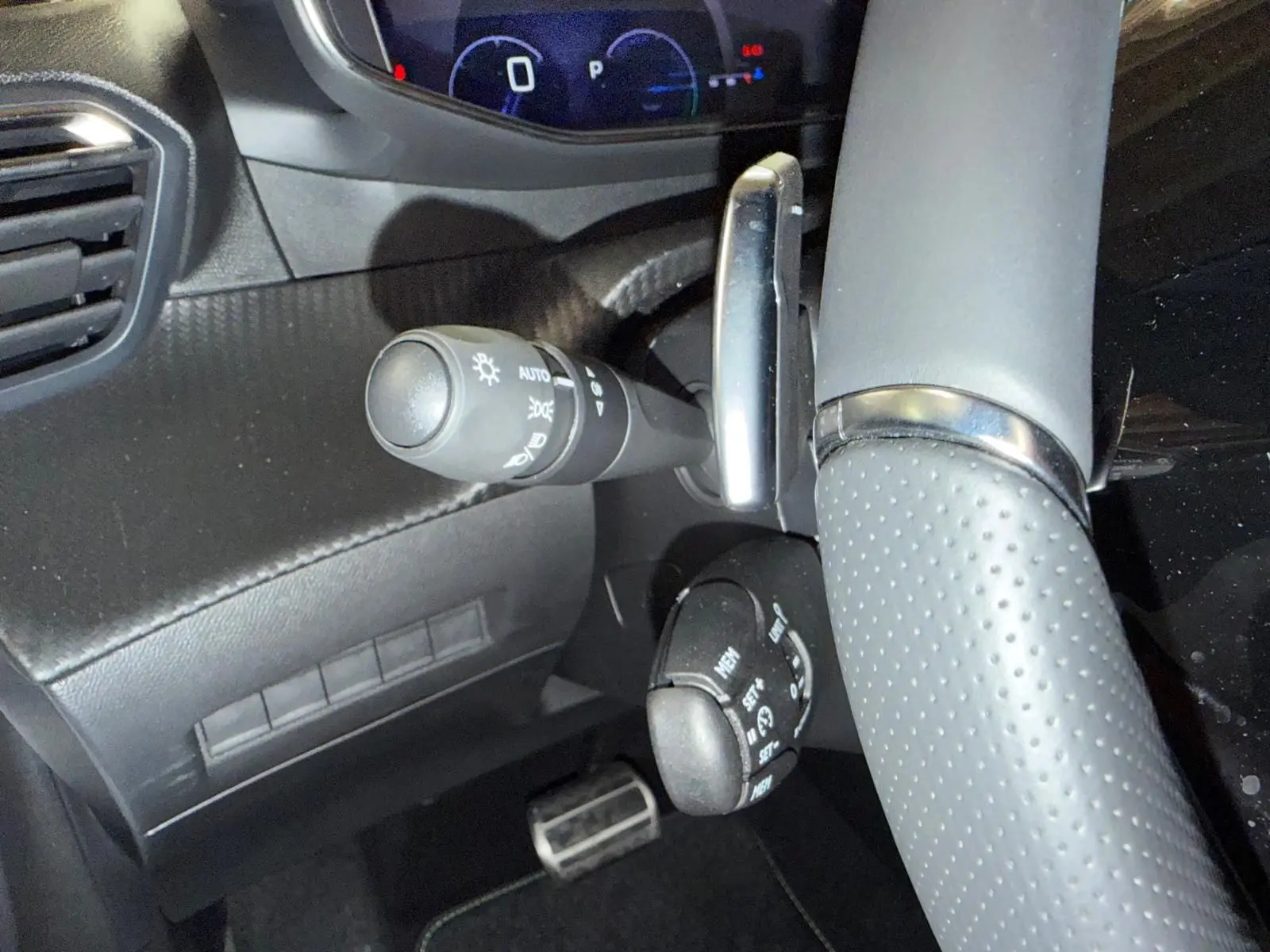 Gros plan sur la partie gauche du volant perforé et les commandes de feux et régulateur d’une Peugeot 208 blanche hybride.