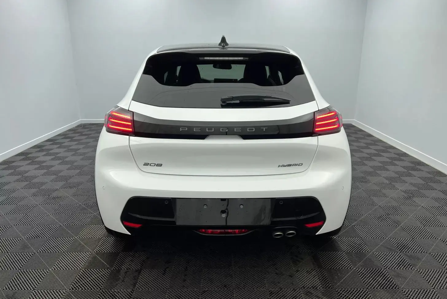 Vue arrière d'une Peugeot 208 blanche hybride 2025 avec feux arrière allumés et double sortie d'échappement côté droit.