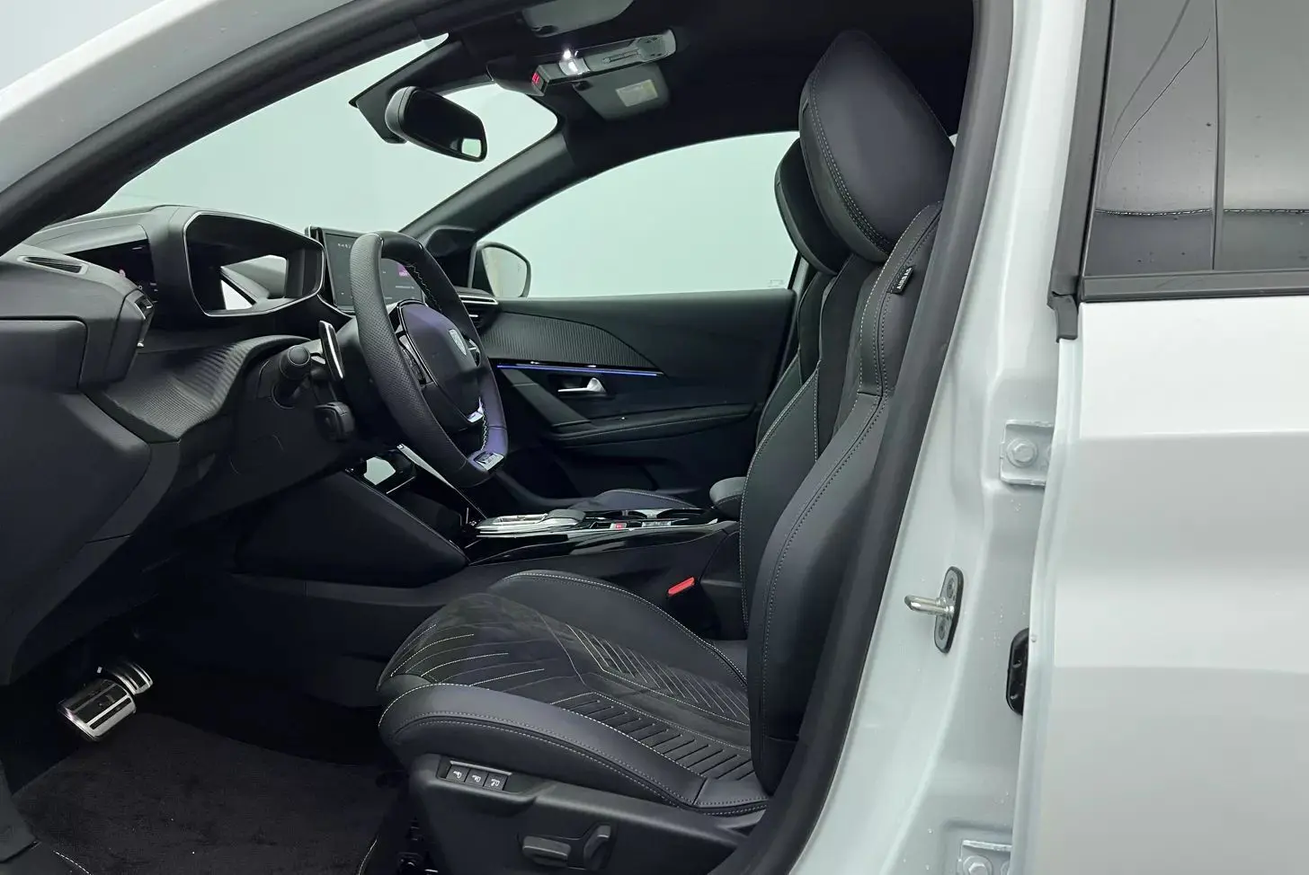 Vue intérieure côté conducteur d'une Peugeot 208 blanche, sièges sport noirs avec surpiqûres et volant multifonction.