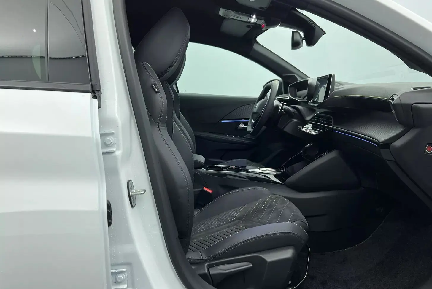 Vue intérieure côté conducteur d'une Peugeot 208 1.2 Hybrid 136 GT blanche, sièges noirs en tissu et tableau de bord moderne.