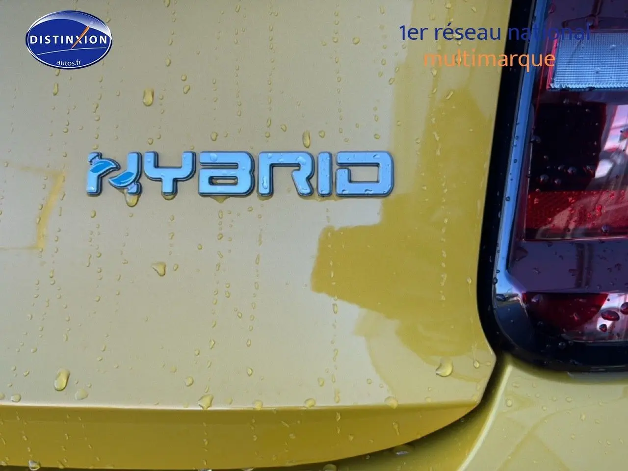 Gros plan sur le logo Hybrid bleu sur la partie arrière droite d'une FIAT 500 Yellow Gold Métal avec gouttes d'eau visibles.