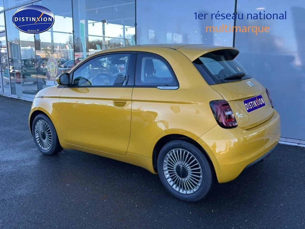 Vue 3/4 arrière droite d'une FIAT 500 Yellow Gold Métal avec jantes alu et becquet arrière, stationnée devant une vitrine.