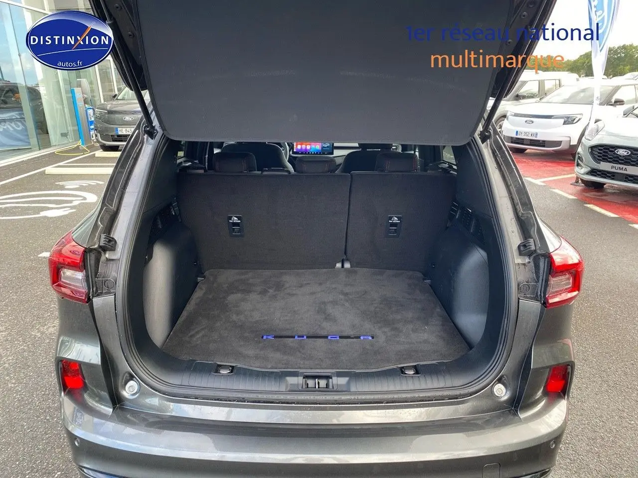 Coffre ouvert en vue arrière d’un Ford Kuga gris Magnetic Fashion Métallisée avec tapis de coffre noir marqué Kuga.
