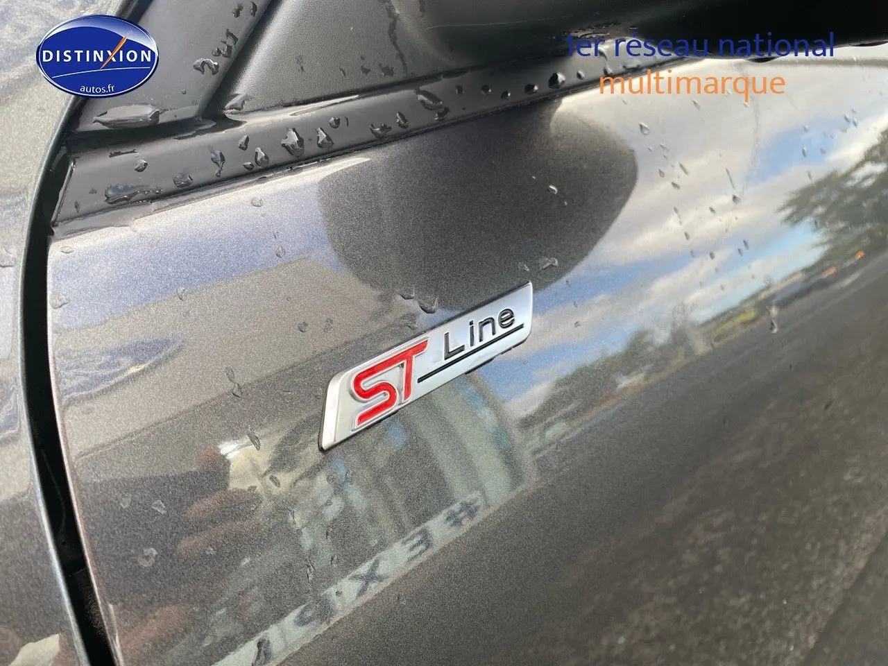 Gros plan sur l'écusson ST-Line rouge et argent sur la porte côté gauche d'un Ford Kuga gris métallisé avec gouttes d'eau.