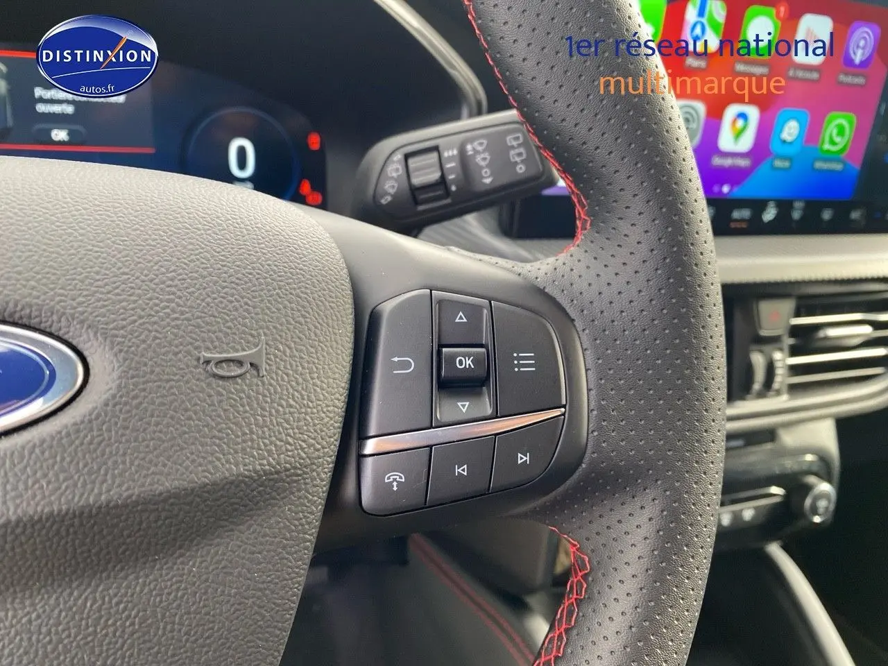 Gros plan sur le volant perforé avec surpiqûres rouges du Ford Kuga 2025, affichage numérique et écran tactile en arrière-plan.