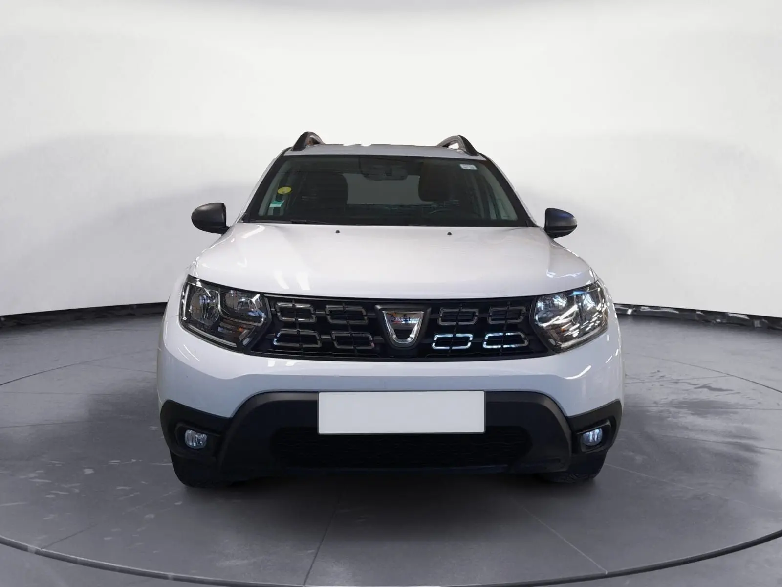 Dacia Duster blanc en 3/4 avant droit, avec calandre noire et jantes alliage 16 pouces, stationné sur un sol gravillonné.