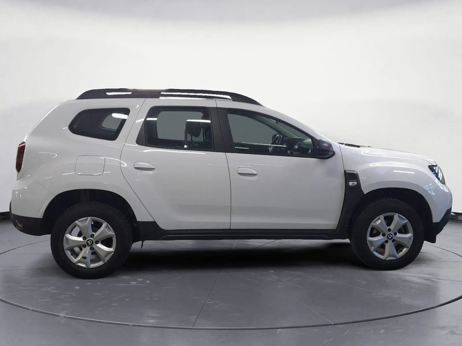 Vue 3/4 arrière droite d'un Dacia Duster blanc 2021 avec jantes alliage et barres de toit sous un abri.